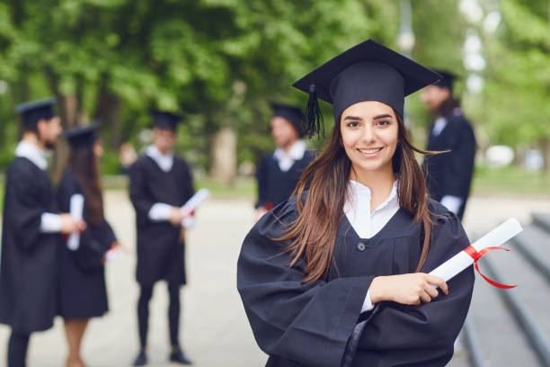 Smiling UK graduate holding a diploma in cap and gown, with other graduates in the background
