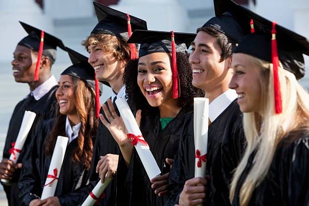 Row of smiling UK graduates in caps and gowns holding diplomas